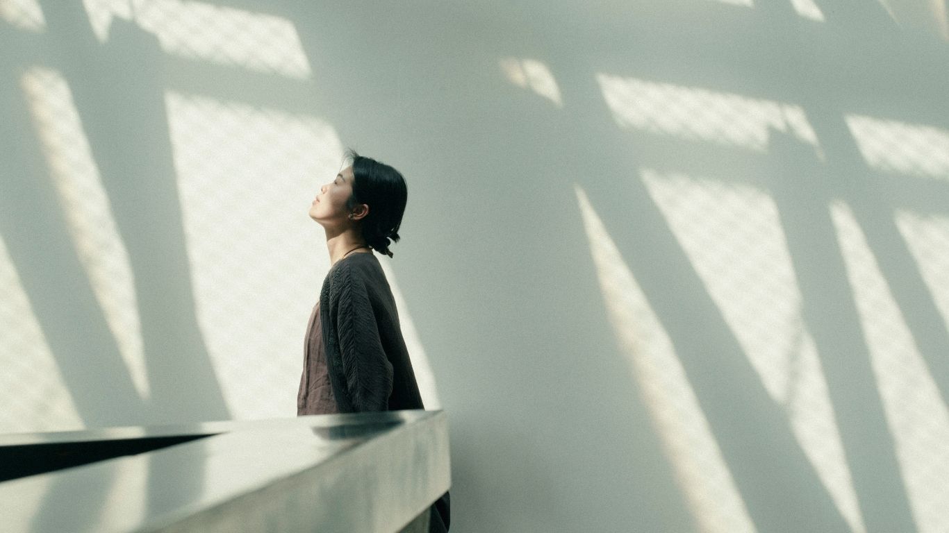 Asiatische Geschäftsfrau im grauen Business-Anzug steht mit geschlossenen Augen in einem lichtdurchfluteten Firmengebäude, Licht und Schatten der Fenster fallen über den Raum – Symbol für inneren Führungszustand und bewusste Präsenz im Leadership.