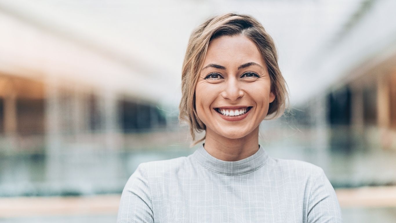 Blonde Führungskraft lächelt selbstbewusst in modernem Business-Umfeld, unscharfer Firmenkorridor im Hintergrund – Symbol für verkörperte digitale Leadership und innere Souveränität.